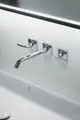 Wand-Waschtischarmatur mit zwei Hebelgriffen und einem langen Auslauf in verchromter Ausführung, montiert an einer weißen Wand mit Waschbecken. Hersteller Ideal Standard.